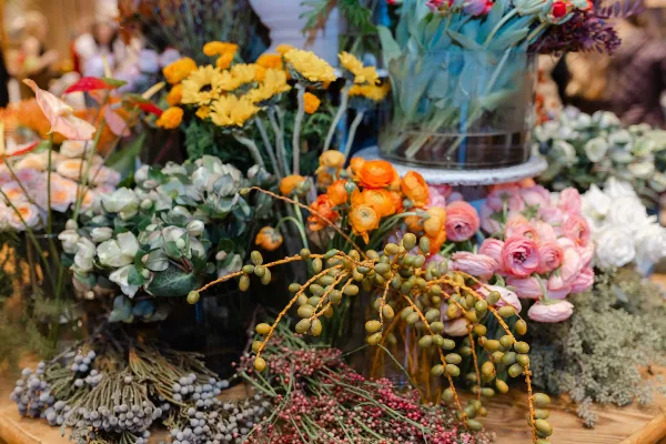 A close-up view of a flower shop display featuring orange ranunculus, pink roses, yellow chrysanthemums, and various greenery arranged in vases.