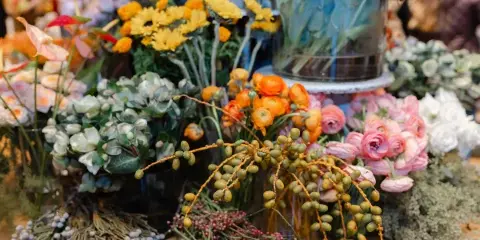 A close-up view of a flower shop display featuring orange ranunculus, pink roses, yellow chrysanthemums, and various greenery arranged in vases.