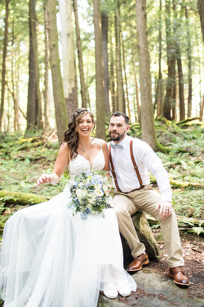 A bride and groom sitting on a log in a forest, with the bride holding a bouquet of white and blue flowers.