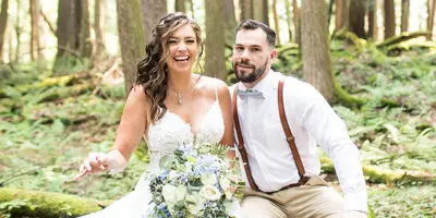 A bride and groom sitting on a log in a forest, with the bride holding a bouquet of white and blue flowers.