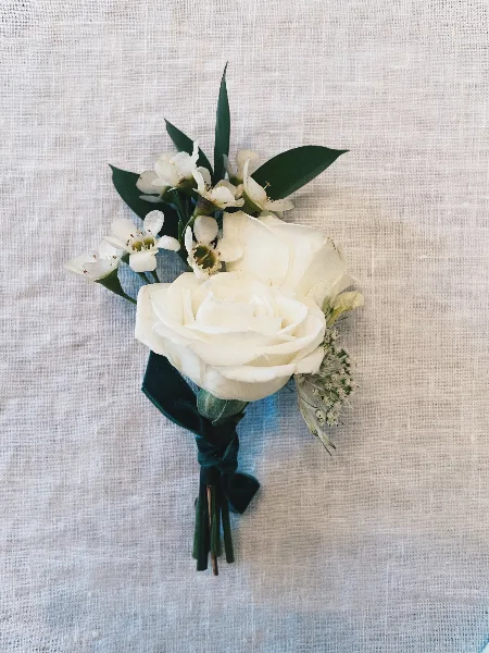 A white rose boutonniere with small white flowers and green leaves tied with a dark green ribbon, resting on a textured white fabric.