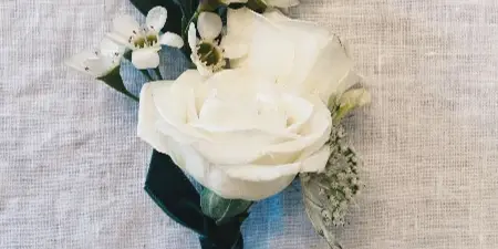 A white rose boutonniere with small white flowers and green leaves tied with a dark green ribbon, resting on a textured white fabric.