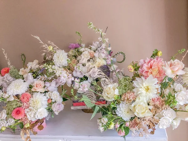 A large, elegant floral arrangement with white and pink roses, dahlias, and dried grasses arranged on a white surface.