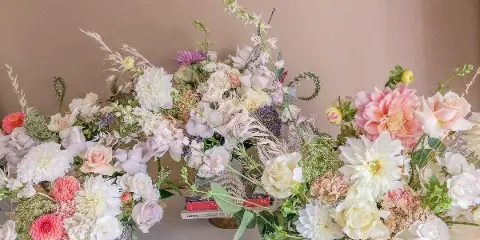 A large, elegant floral arrangement with white and pink roses, dahlias, and dried grasses arranged on a white surface.