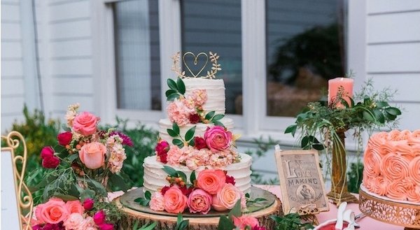 A multi-tiered white wedding cake decorated with pink and red roses, greenery, and a gold heart topper, displayed on a wooden stand.