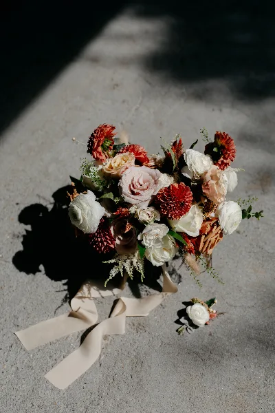 A wedding bouquet with red dahlias, roses, and ranunculus, with a matching boutonniere, resting on a concrete surface.