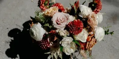 A wedding bouquet with red dahlias, roses, and ranunculus, with a matching boutonniere, resting on a concrete surface.
