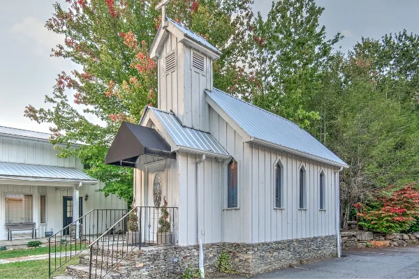 Exterior view of a small white wooden chapel with a steeple and metal roof, surrounded by trees.