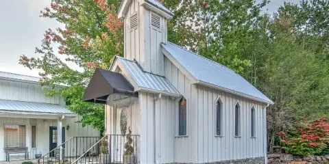 Exterior view of a small white wooden chapel with a steeple and metal roof, surrounded by trees.