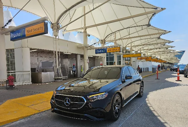 A black Mercedes-Benz luxury sedan parked at an airport curb near a TSA PreCheck security lane.