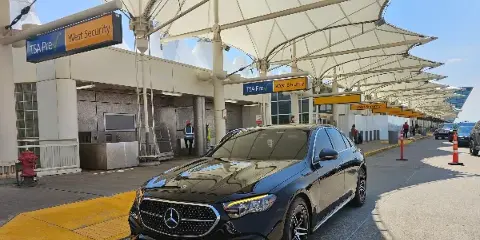 A black Mercedes-Benz luxury sedan parked at an airport curb near a TSA PreCheck security lane.