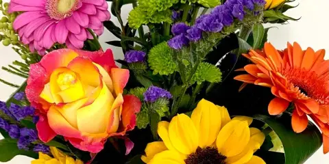 A colorful floral arrangement featuring sunflowers, pink and orange gerbera daisies, a rose, and greenery.