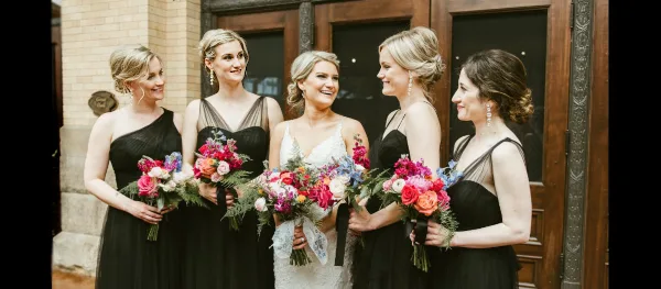 A bride and four bridesmaids standing together outside a venue, all with styled hair and makeup, holding bouquets.