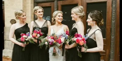 A bride and four bridesmaids standing together outside a venue, all with styled hair and makeup, holding bouquets.