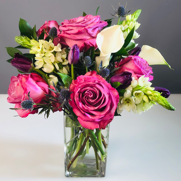 A vibrant floral arrangement with pink roses, white calla lilies, and purple accents in a clear glass vase.