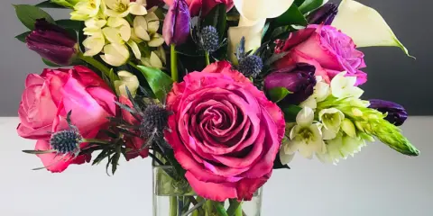A vibrant floral arrangement with pink roses, white calla lilies, and purple accents in a clear glass vase.