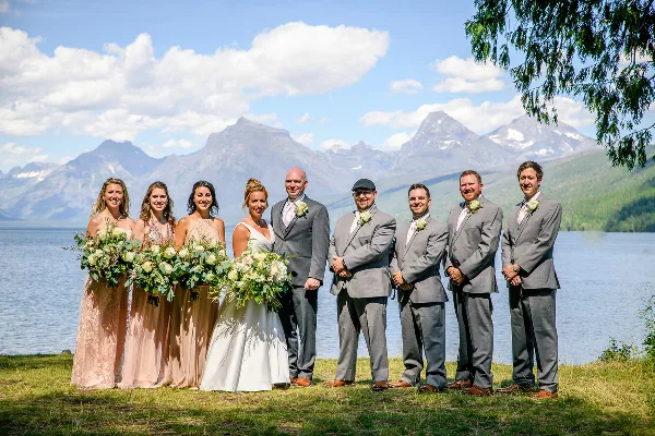 A wedding party posing outdoors with a lake and mountain peaks in the background.