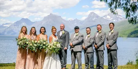 A wedding party posing outdoors with a lake and mountain peaks in the background.