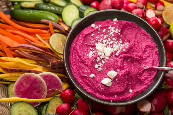 A bowl of vibrant pink beet hummus topped with crumbled white cheese, surrounded by an assortment of fresh vegetable sticks including carrots, cucumbers, and radishes.