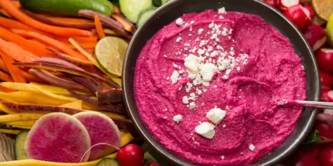 A bowl of vibrant pink beet hummus topped with crumbled white cheese, surrounded by an assortment of fresh vegetable sticks including carrots, cucumbers, and radishes.