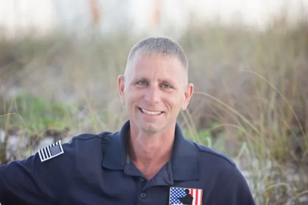 A professional portrait of a smiling man in a dark blue uniform with an American flag patch, standing outdoors against a blurred background of tall grass.