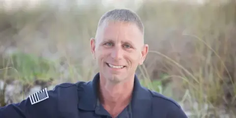 A professional portrait of a smiling man in a dark blue uniform with an American flag patch, standing outdoors against a blurred background of tall grass.