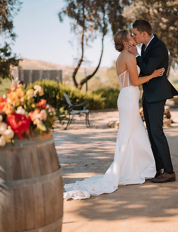 A bride and groom kissing outdoors next to a wine barrel during a wedding ceremony.