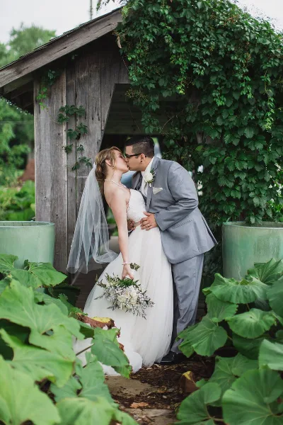 Bride and groom kissing under a vine-covered wooden structure, with the bride holding a white and green floral bouquet.