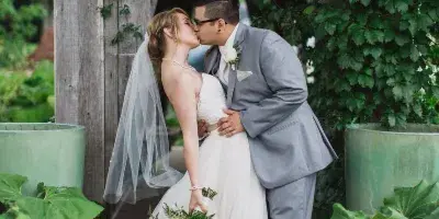 Bride and groom kissing under a vine-covered wooden structure, with the bride holding a white and green floral bouquet.