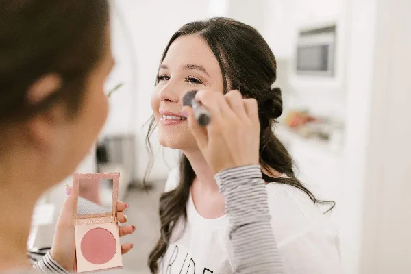 A makeup artist applies blush to a smiling client using a brush and compact mirror.