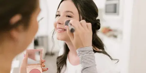 A makeup artist applies blush to a smiling client using a brush and compact mirror.