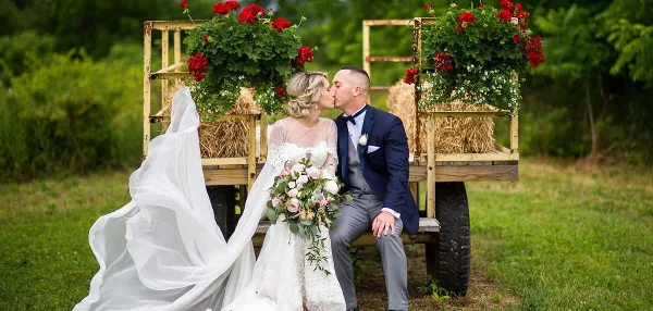 A bride and groom kissing while seated on a rustic wooden wagon decorated with hay bales and red flowers in a grassy field.