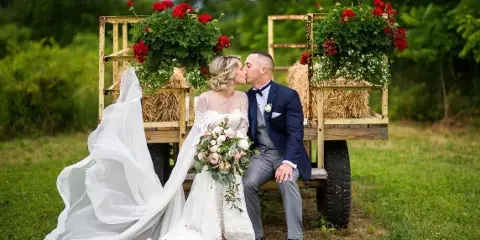 A bride and groom kissing while seated on a rustic wooden wagon decorated with hay bales and red flowers in a grassy field.