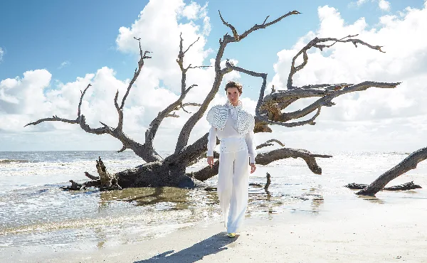 A model with styled hair and makeup wearing a white outfit walks on a beach in front of a large driftwood tree.