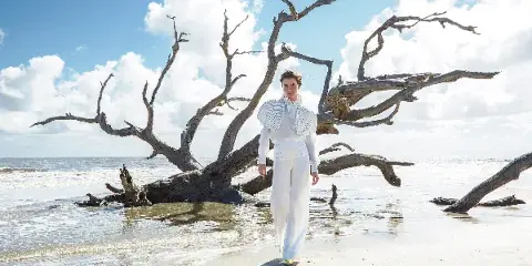A model with styled hair and makeup wearing a white outfit walks on a beach in front of a large driftwood tree.