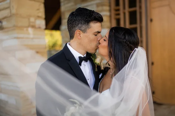 A bride and groom sharing a kiss in front of a stone building, with the bride's veil flowing in the foreground.