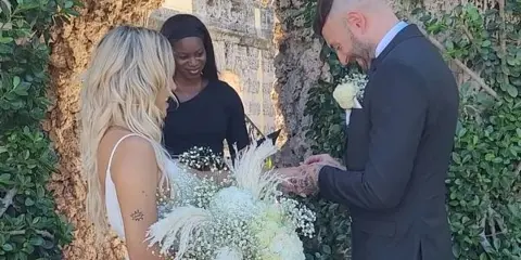 A wedding officiant in a black dress stands between a bride and groom during an outdoor ceremony.
