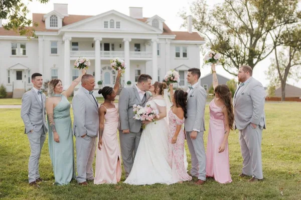 A wedding party posing on the lawn in front of a large white historic mansion with a columned entrance.