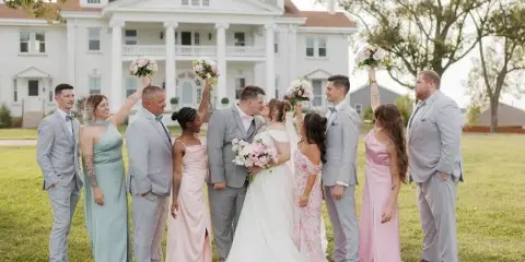 A wedding party posing on the lawn in front of a large white historic mansion with a columned entrance.