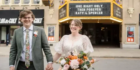 A bride and groom holding hands in front of the Michigan Theater marquee, which displays their names and the text 'One Night Only Then Forever'.
