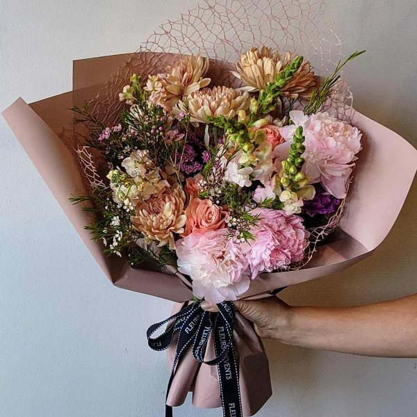 A hand holding a wrapped bouquet of pink and peach flowers with a branded ribbon