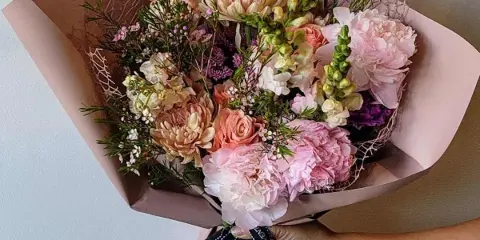 A hand holding a wrapped bouquet of pink and peach flowers with a branded ribbon