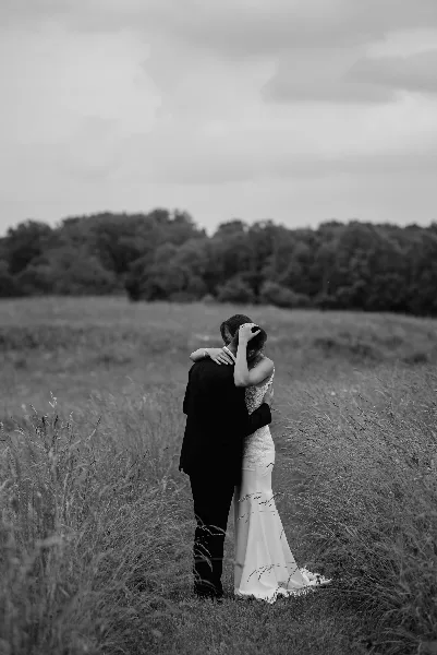 A black and white photograph of a bride and groom embracing in a grassy field.