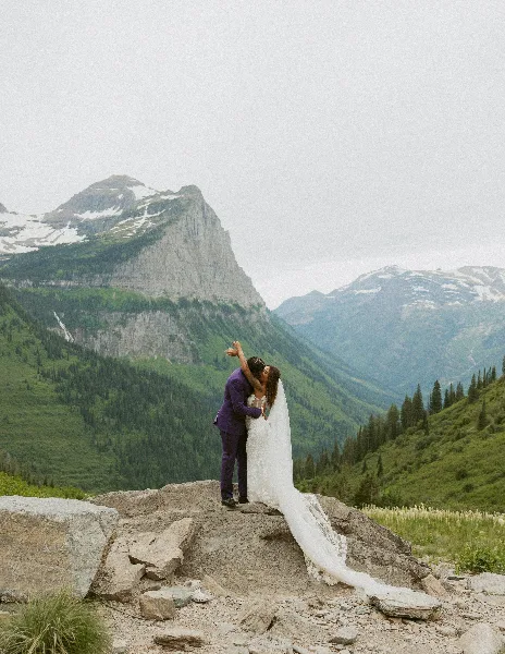 A bride and groom embracing on a rocky outcrop with a backdrop of green mountains and snow-capped peaks.