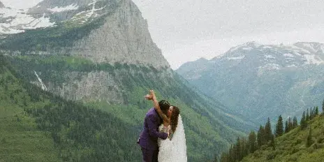 A bride and groom embracing on a rocky outcrop with a backdrop of green mountains and snow-capped peaks.