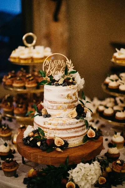 A two-tier semi-naked wedding cake decorated with gold leaf, fresh figs, and berries, displayed on a wooden stand surrounded by cupcakes.