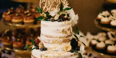 A two-tier semi-naked wedding cake decorated with gold leaf, fresh figs, and berries, displayed on a wooden stand surrounded by cupcakes.