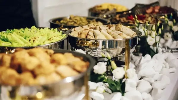 A catering buffet table featuring multiple stainless steel chafing dishes filled with appetizers and salads, decorated with white flowers and white stones.