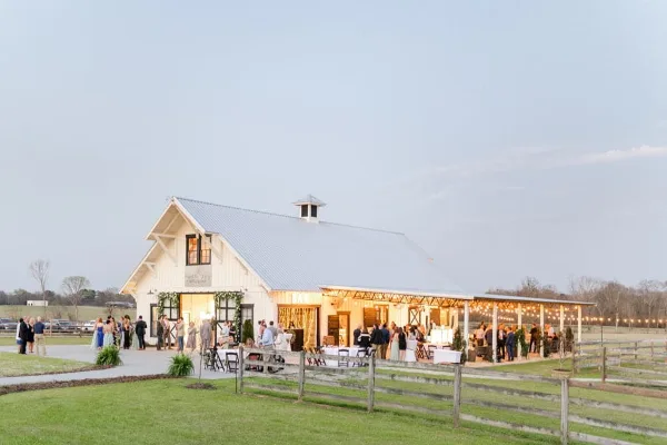 A wide shot of a white barn event venue with a covered patio, string lights, and guests gathered on the lawn at dusk.