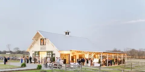 A wide shot of a white barn event venue with a covered patio, string lights, and guests gathered on the lawn at dusk.